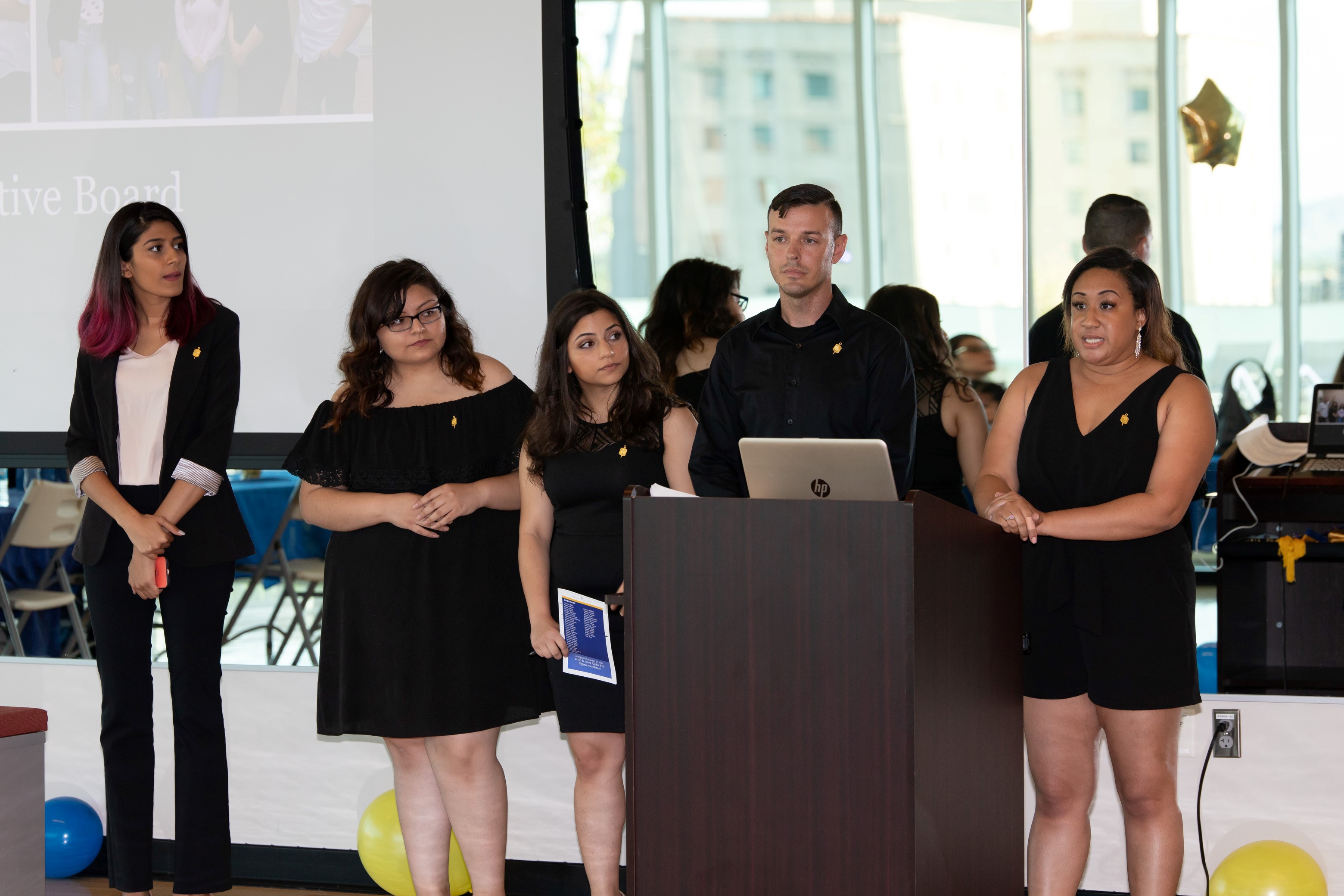 five students in black formal attire speaking at a podium with a computer 