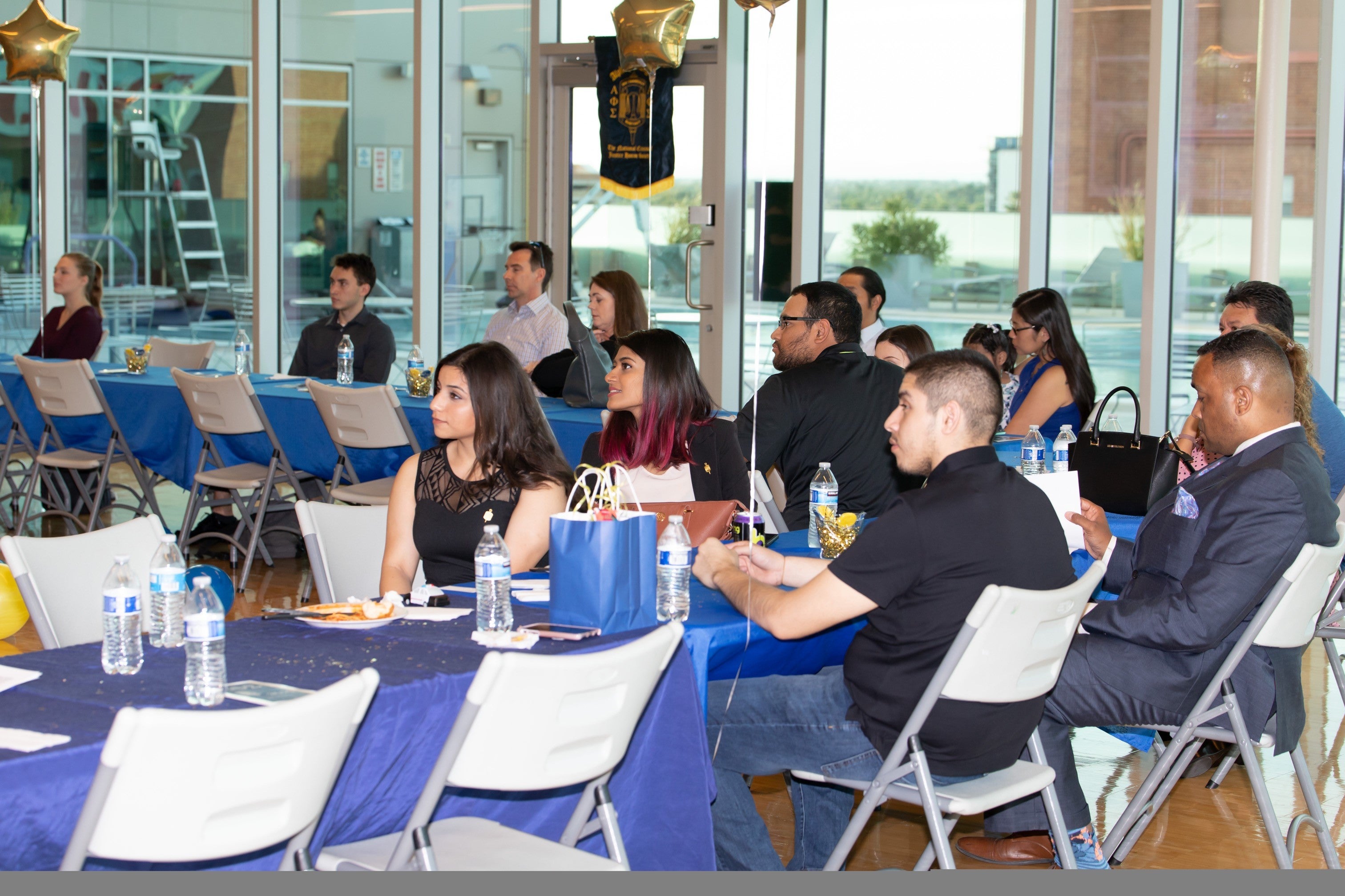 students in black formal attire sitting at a table with a blue tablecloth and refreshments